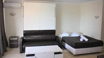 A compact room with a black leather sofa, a bed with a dark blanket, a white bedside table, and a small white cabinet.