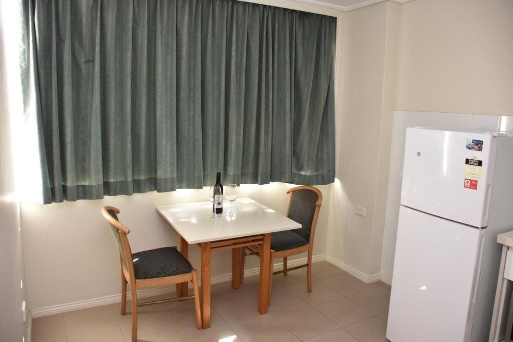 A small dining area with a table and two chairs, a refrigerator, and a window with curtains.