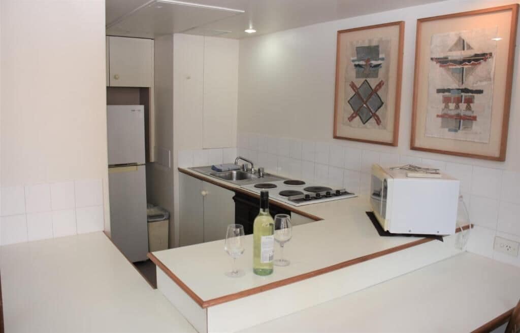 Eine kleine Küche mit weißer Arbeitsplatte, eingebautem Herd, Spüle und Kühlschrank. Über der Arbeitsplatte befinden sich zwei gerahmte Kunstwerke an der Wand.
