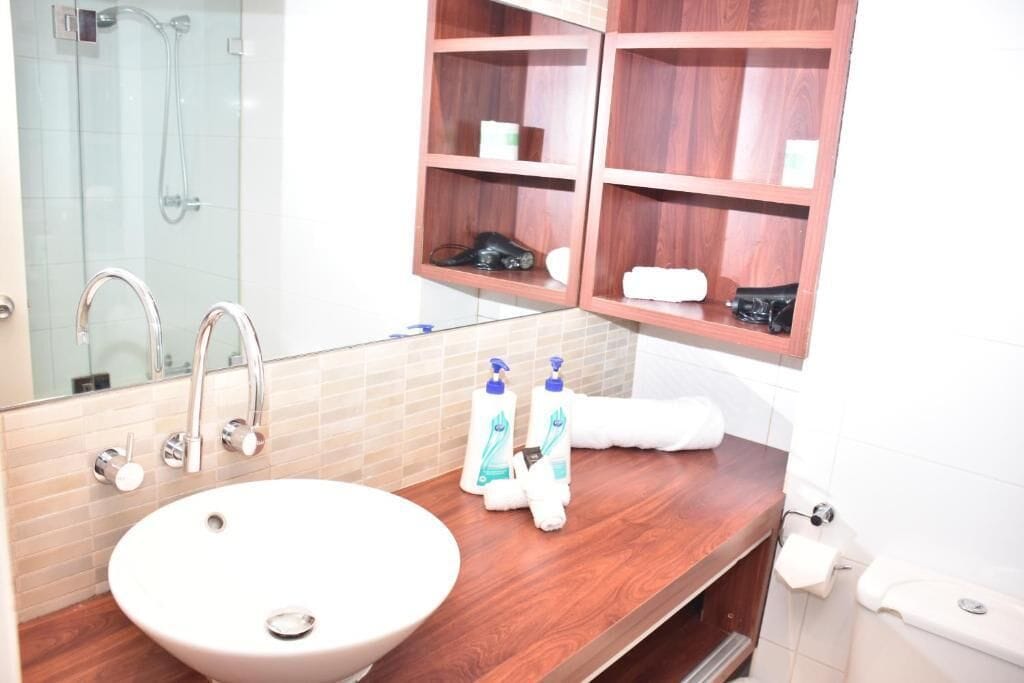 A bathroom with a large sink, a mirror, a shower, and wooden shelves with toiletries.