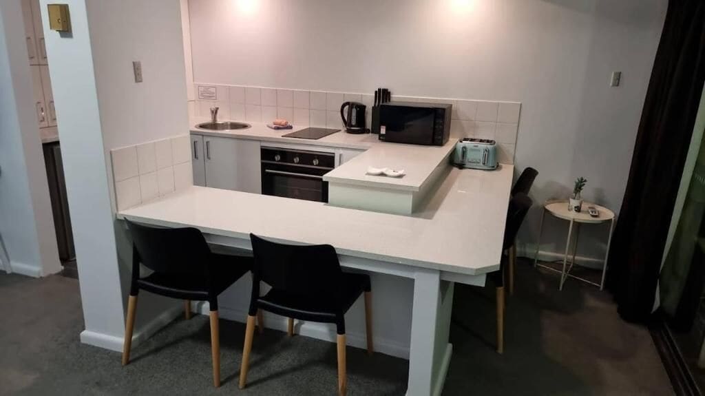 Une cuisine compacte avec un plan de travail blanc, deux chaises noires, un four à micro-ondes, un grille-pain et une petite table ronde agrémentée d’une plante.
