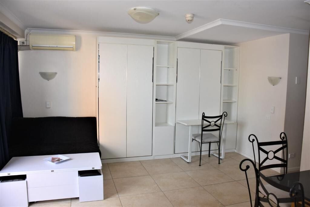 A compact room with a bed, a small table, a chair, and a white wardrobe with shelves.