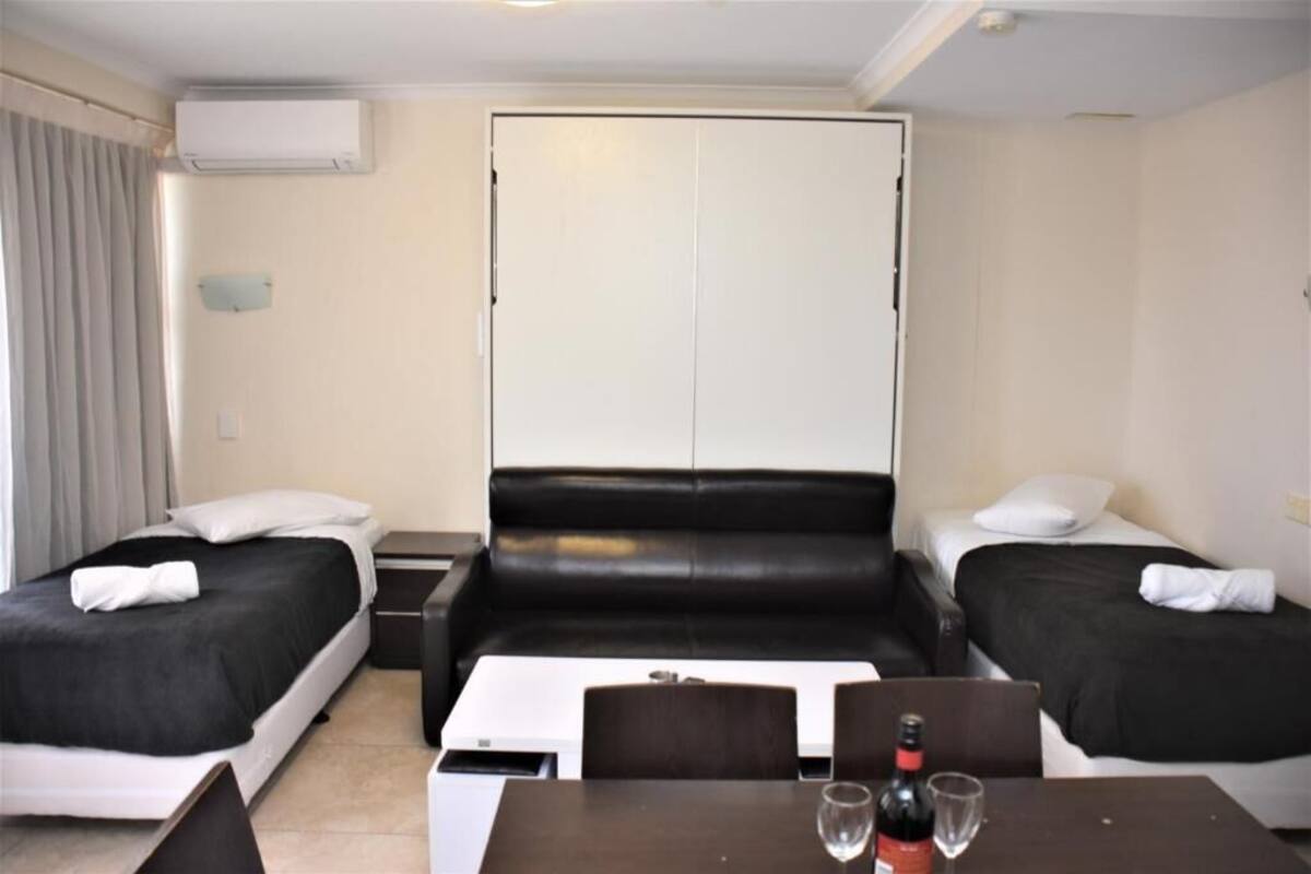 A hotel room with two single beds, a sofa bed, a small table with a bottle and glass, and a white air conditioning unit on the wall.