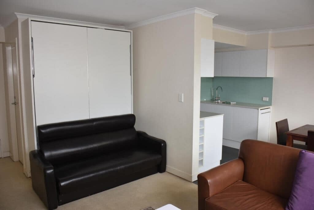A compact living space with a black leather sofa, a brown leather armchair, and a kitchenette area.