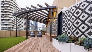 Una terraza en la azotea con suelo de madera, dos sofás grises, una maceta blanca con suculentas y una pared con motivo geométrico.