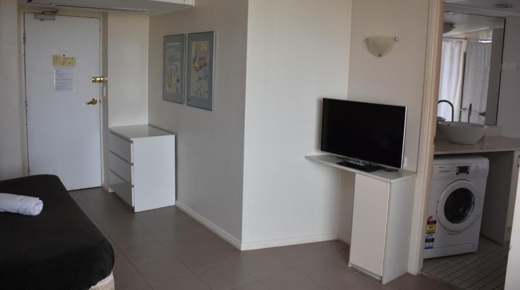 A compact hotel room with a bed, a small desk, a television, a washing machine, and a bathroom with a sink and mirror.