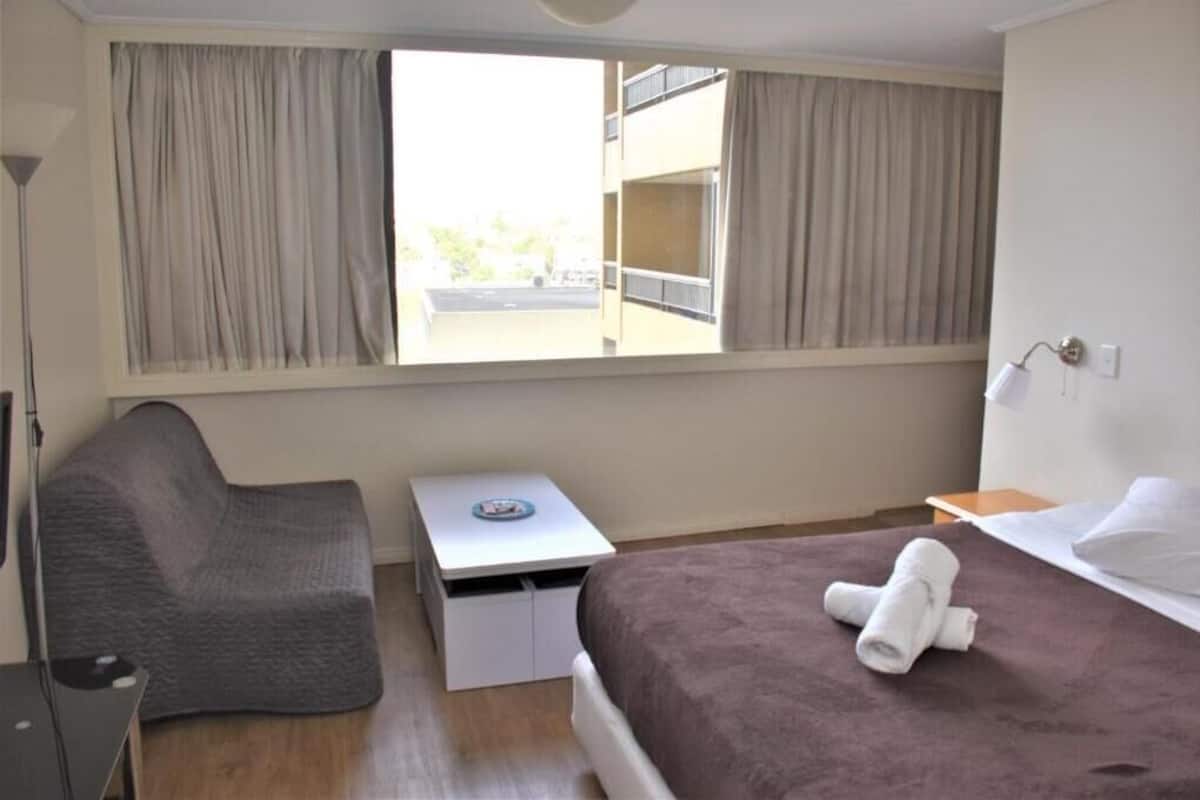 A hotel room with a bed, a small desk, a chair, and a window with curtains.