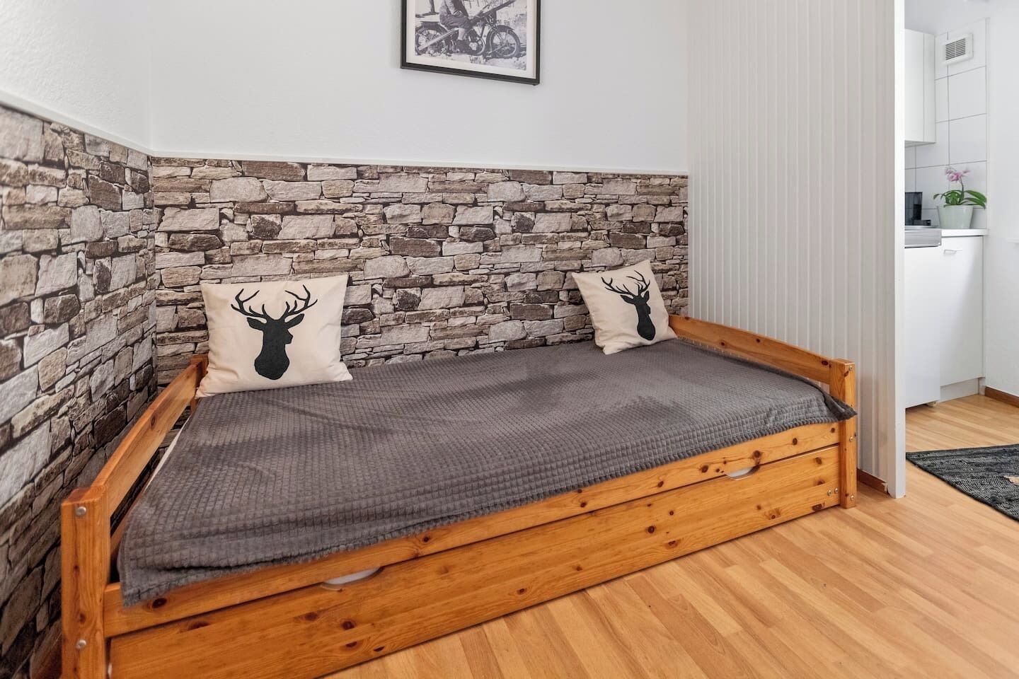 A wooden bed with a stone wall backdrop, two pillows with deer head designs, and a framed picture above the bed.