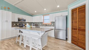 Una cocina con armarios blancos, una isla central con taburetes, un refrigerador de acero inoxidable y una puerta de madera.