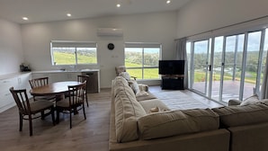 A spacious living area with a large sofa, dining table, and chairs. There is a television and a ceiling fan. The room has large windows offering a view of the outdoors. - Cosy and comfortable executive Unit with gorgeous views and close to town centre (Clare)