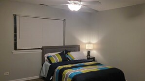 Un dormitorio con cama, ventilador de techo, ventana con persianas y mesita de noche con lámpara.