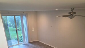 Amplia habitación con piso de madera, ventilador de techo y puerta corrediza de vidrio que da a un jardín.