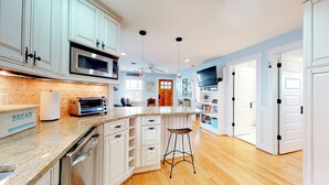 A modern kitchen with white cabinetry, a granite countertop, a built-in oven, and a large island with stools. - Charming Beach Cottage – Walk to Ocean, Relaxing Porch & Private Parking (Carolina Beach)