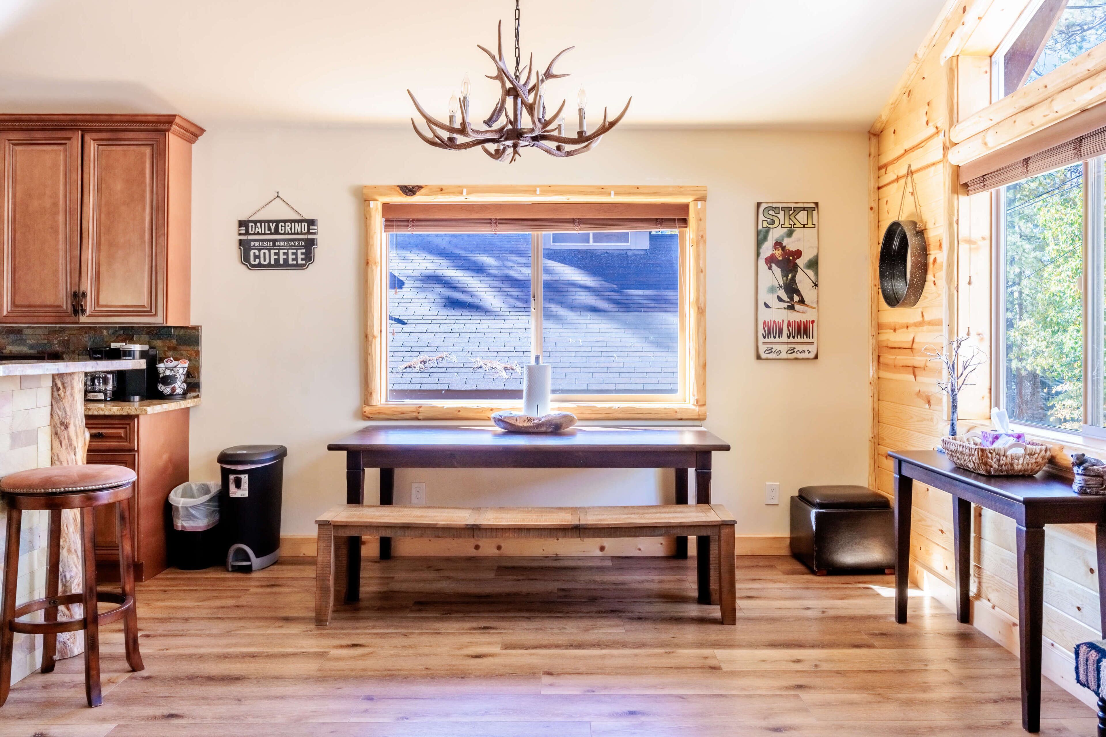 A kitchen with wooden cabinets, a dining table, a bench, a chandelier, and a window with a view of a brick building.