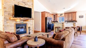 A modern living room with a fireplace, a large flat-screen TV mounted on a brick wall, a brown leather sofa, and a wooden coffee table. - Bearadise ~ Enchanted Dream ~ Mountain Escape (Big Bear Lake)