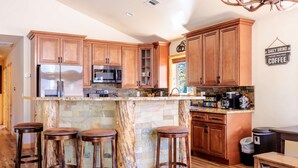 A kitchen with wooden cabinets, a granite countertop, a stainless steel refrigerator, a microwave, and a coffee machine. There are four wooden stools and a trash can. - Bearadise ~ Enchanted Dream ~ Mountain Escape (Big Bear Lake)