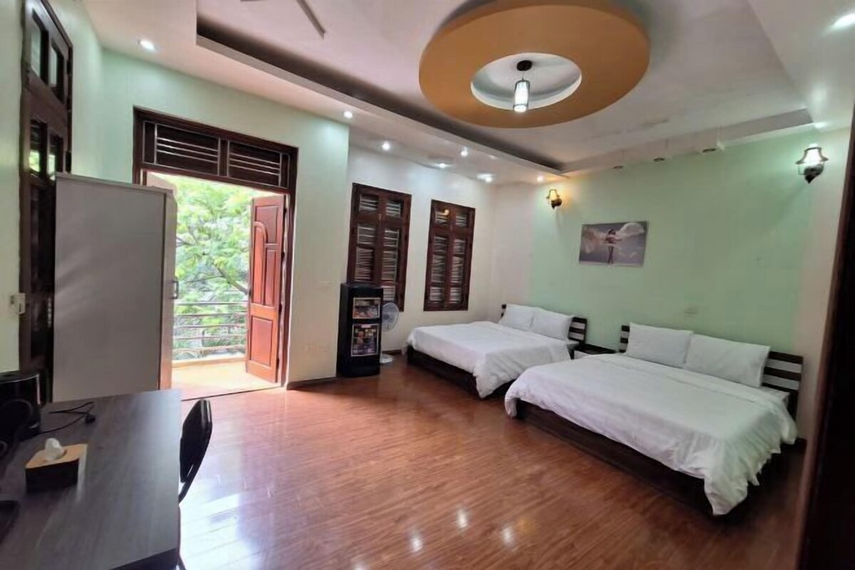 A hotel room with two beds, a dining table, a refrigerator, and a view of greenery outside.