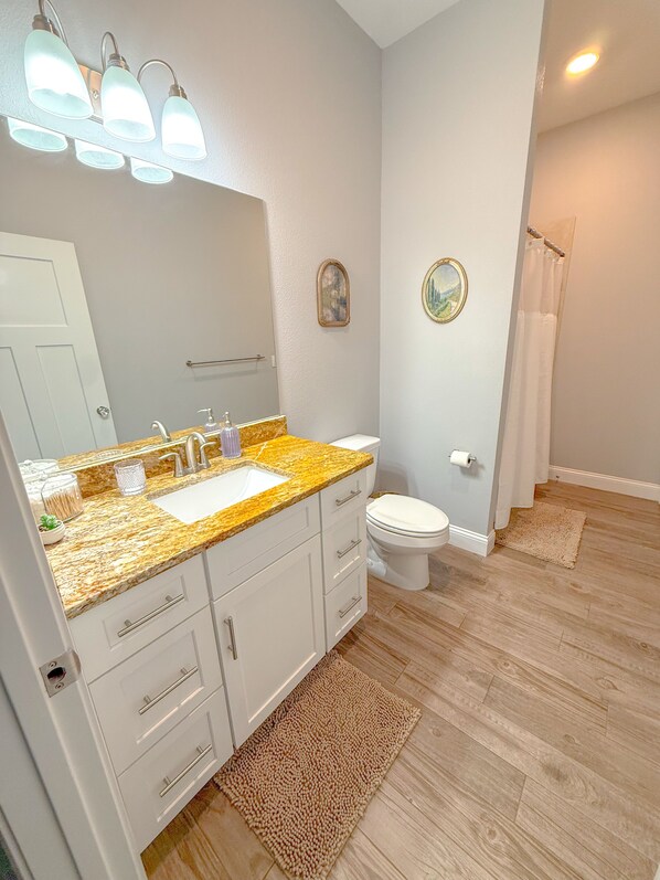 Un baño con encimera de granito, armarios blancos, inodoro y cortina de ducha.