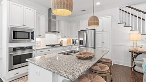 A modern kitchen with white cabinetry, stainless steel appliances, a granite countertop, and a central island with stools. - Magnolia Mint | Stylish 4BR Retreat w/ Golf Cart, Bikes, Sleeps 10, Resort Amenities (Santa Rosa Beach)