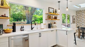Une cuisine moderne avec des meubles blancs, une grande fenêtre donnant sur un espace verdoyant et des luminaires suspendus.