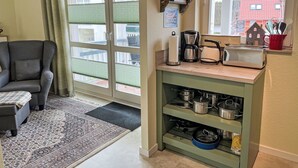 Una sala de estar acogedora con un sillón gris, un tapete estampado y una zona de cocina con cafetera, tostadora y estantes con ollas.