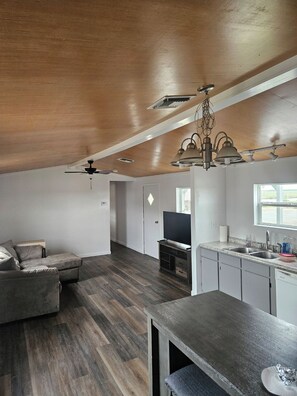 A spacious living area with a wooden ceiling, a dining table, a kitchen with white cabinets, and a television.