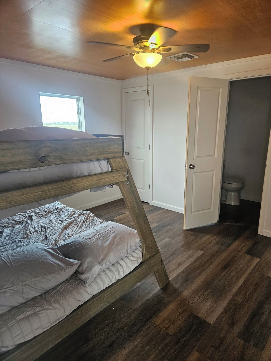 A room with a bunk bed, wooden flooring, a ceiling fan, a window, and a door leading to a bathroom.