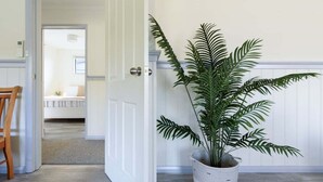 Un couloir avec une porte blanche donnant sur une chambre. Une grande plante en pot est placée dans un coin.