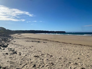 Una playa arenosa con huellas, cielo despejado y una línea de costa distante con árboles.