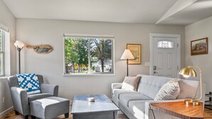 A living room with a grey sofa, two armchairs, a coffee table, and a lamp. There is a window with a view of trees and a door in the background. - The Cottage on Cherry Lane (Spokane)