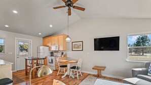 A spacious kitchen with wooden cabinets, a dining area with a table and chairs, a ceiling fan, and a large window with a view of trees. - The Cottage on Cherry Lane (Spokane)