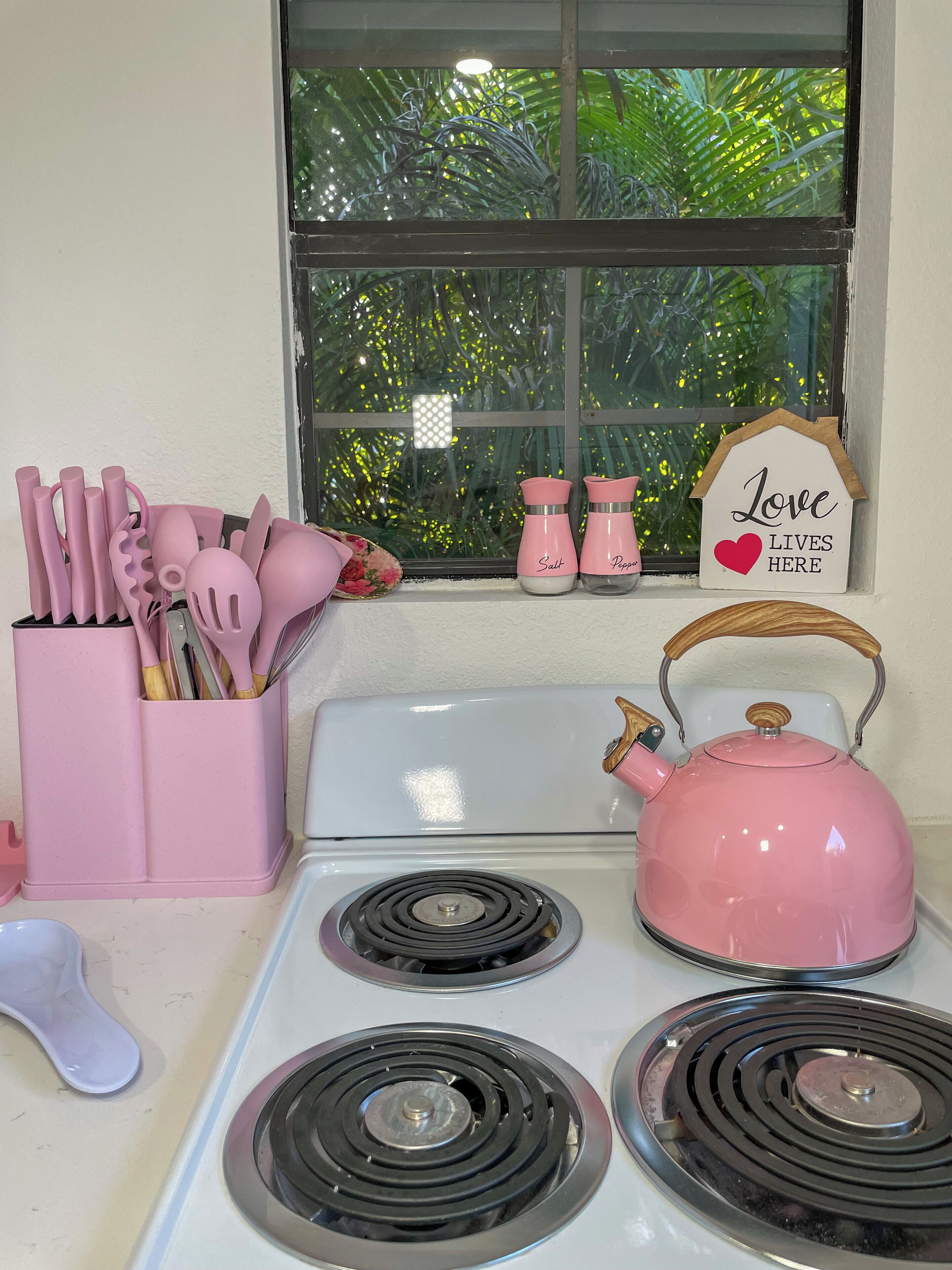 Una cucina con utensili rosa, un bollitore rosa e una finestra con un cartello che dice 'Love Lives Here'.