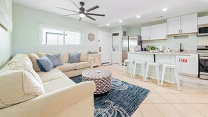 A modern living room with a sofa, ottoman, and a ceiling fan. It includes a kitchen with white cabinets, a refrigerator, and a microwave. - Seaside Sanctuary • 2 Min Walk to Okaloosa Beach (Fort Walton Beach)