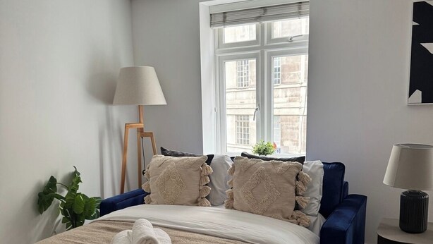Un dormitorio con una cama grande, un sofá azul, una ventana con persianas, una lámpara y una planta.