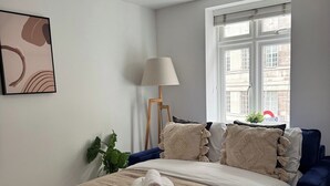 Una cama bien hecha con una manta color beige y toallas enrolladas, un sillón azul, una lámpara de pie y una ventana con vistas a edificios.