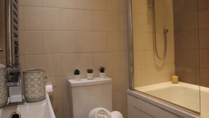 Un baño con inodoro, lavamanos y ducha. Hay tres pequeñas macetas sobre el tanque del inodoro, una toalla en el suelo y una botella de jabón en la bañera.