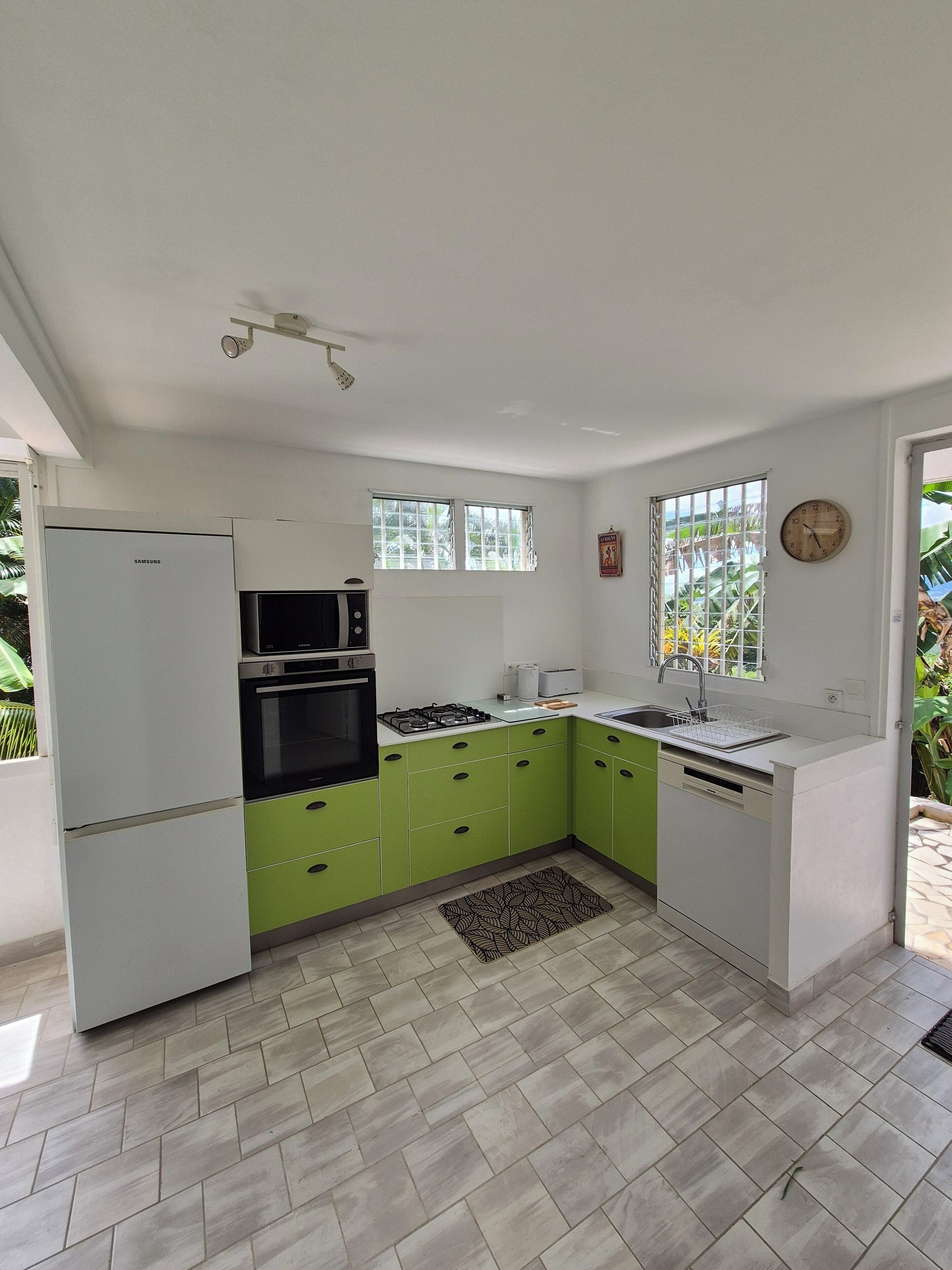 A modern kitchen with white and green cabinets, a built-in oven, microwave, and gas stove. There is a window with a view of greenery outside, and a clock on the wall.