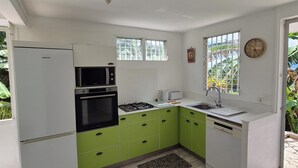 Una cocina moderna con armarios blancos y verdes, horno empotrado, microondas y estufa a gas. Hay una ventana con vista a zonas verdes, y un reloj en la pared.