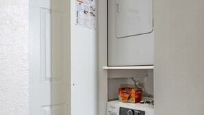 A compact laundry room with a front-loading washer and dryer, a small shelf with detergent boxes, and a door with a maintenance poster. - 3-Min Walk to Ski Lift | Mountain-View Chalet (Angel Fire)