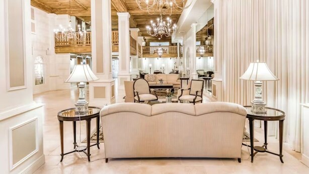 A luxurious hotel lobby with a large beige sofa, two side tables with lamps, a chandelier, and a dining area in the background.