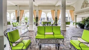 A spacious outdoor seating area with green cushioned chairs and tables, surrounded by columns and potted plants.