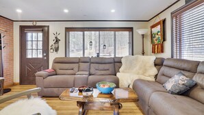 A living room with a brown leather sofa, a wooden coffee table with a bowl and remote controls, a fluffy white throw, and a door with a wreath.