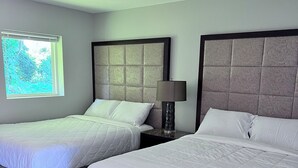 A hotel room with two beds, a nightstand with a lamp, and a window with a view of greenery. - Your home away from home (Rocky Mount)