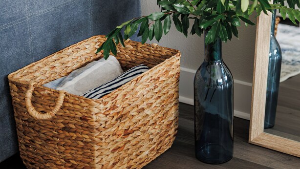 Een rieten mand met een kussen, een donkerblauwe vaas met een plant, en een spiegel met houten lijst op een houten vloer.