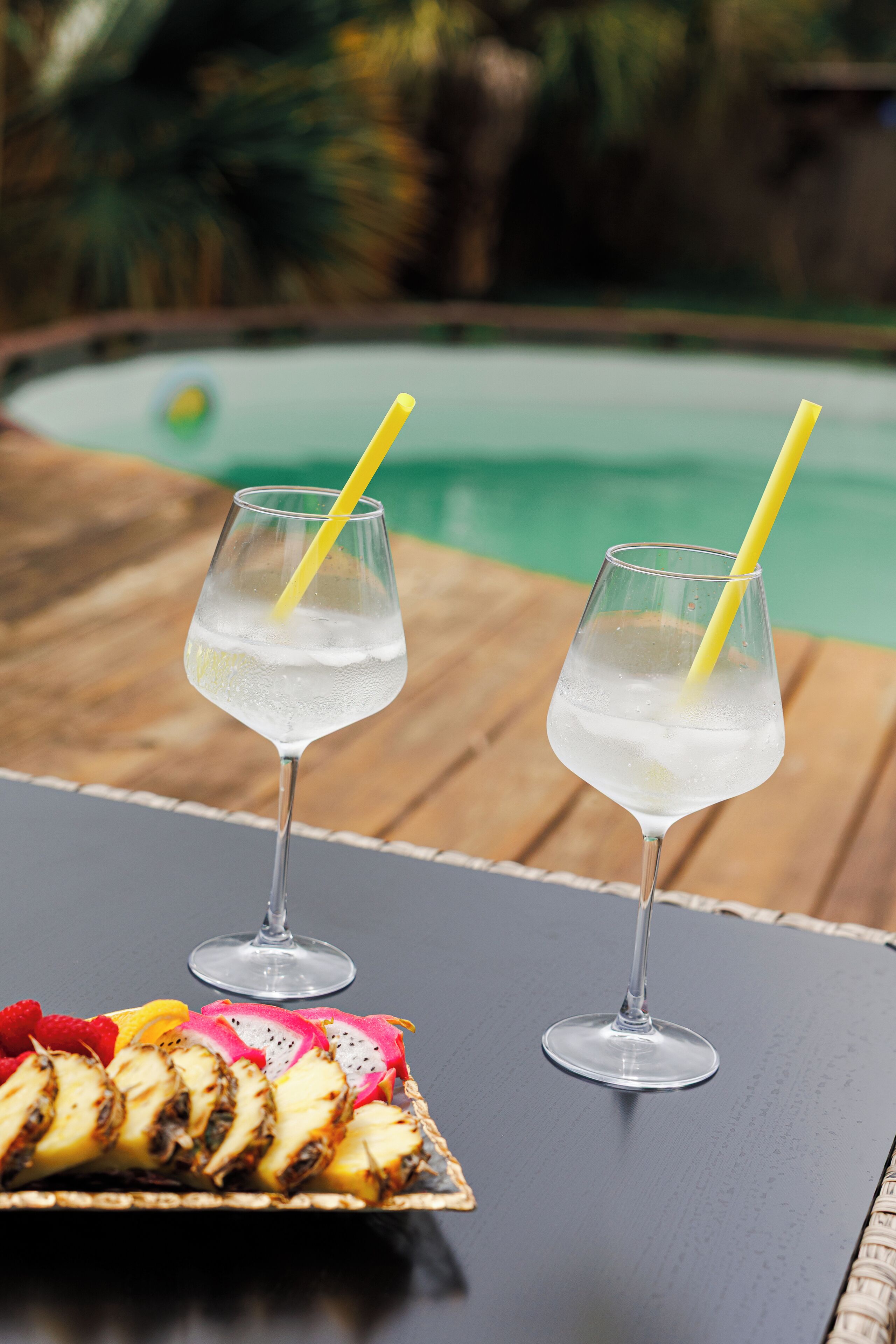 Two glasses of a clear beverage with a yellow straw, a plate of sliced fruit, and a pool in the background.