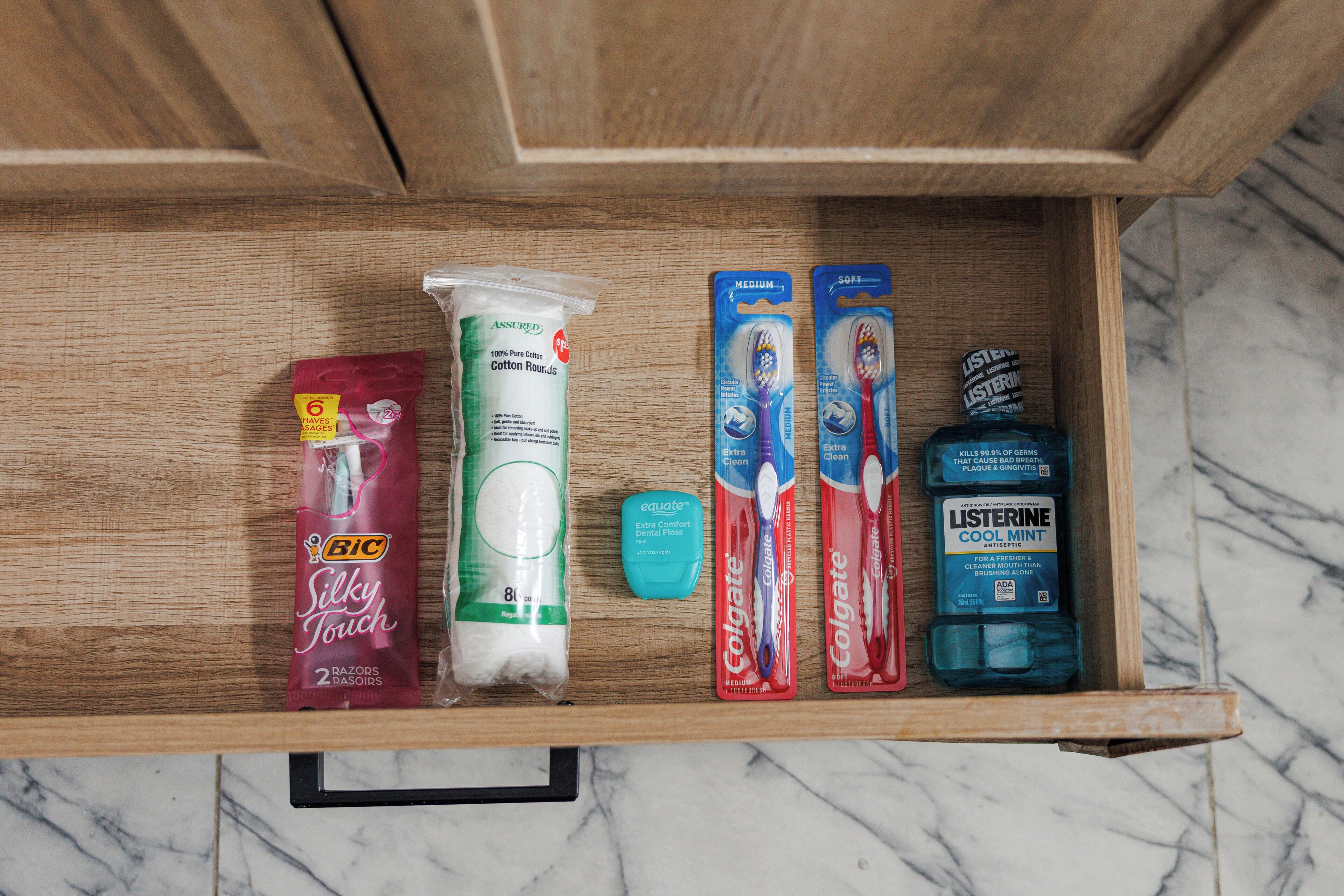 Een lade met diverse toiletartikelen, waaronder een pak Bic Silky Touch scheermesjes, een tube Assured Cotton Rounds, Colgate tandpasta en een fles Listerine Cool Mint mondwater.