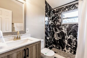 Une salle de bain avec un mur de douche en marbre, un lavabo blanc avec un robinet doré et des toilettes avec une étiquette verte.