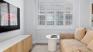 Ein Wohnzimmer mit einem an der Wand montierten Flachbildfernseher, einem weißen Sofa, einem kleinen weißen Beistelltisch und einem Fenster mit Jalousien.