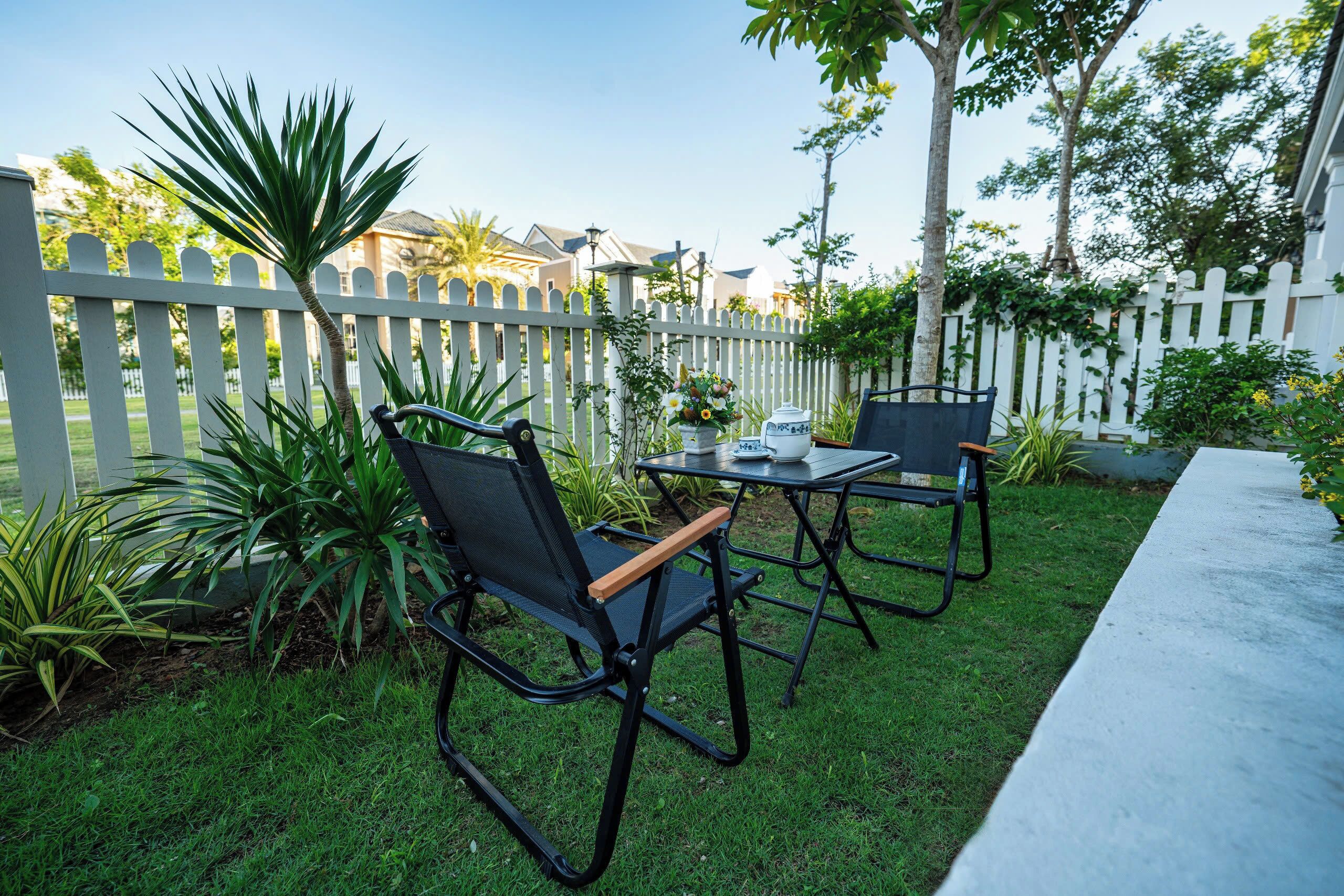 An outdoor patio area with two black chairs and a table, surrounded by a white picket fence and greenery.