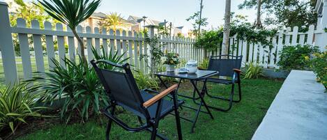 Un patio exterior con dos sillas negras y una mesa, rodeado por una cerca de piquetes blancos y follaje.